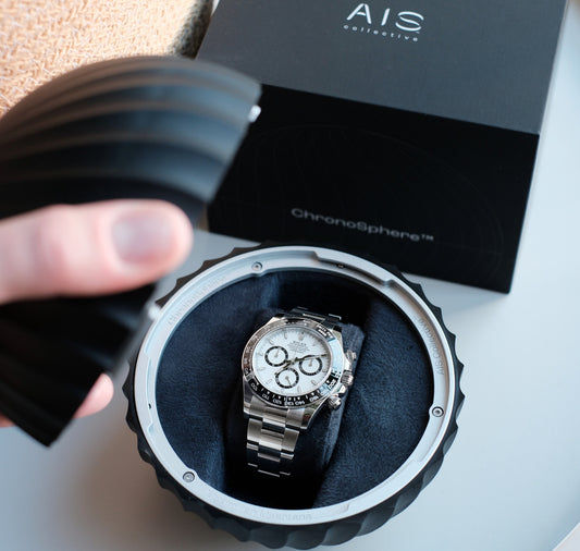 A person is opening a round, black, ridged ChronoSphere to reveal a Rolex Daytona stainless steel chronograph watch with a white dial and black subdials nestled inside a black velvet cushion. A black box in the background is partially visible.