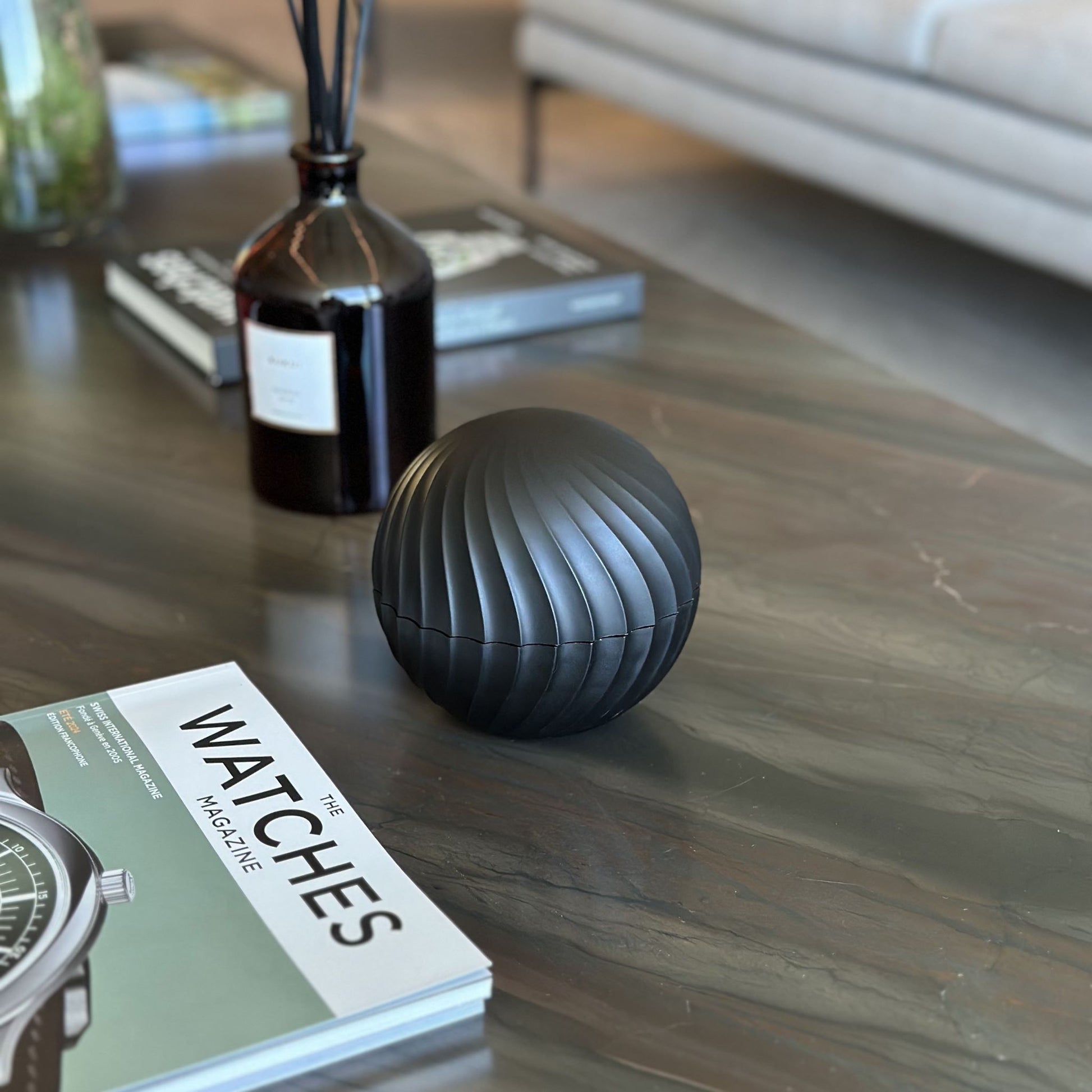 The matte black ChronoSphere luxury watch storage case styled as a minimalist design object on a dark marble coffee table next to The Watches Magazine.
