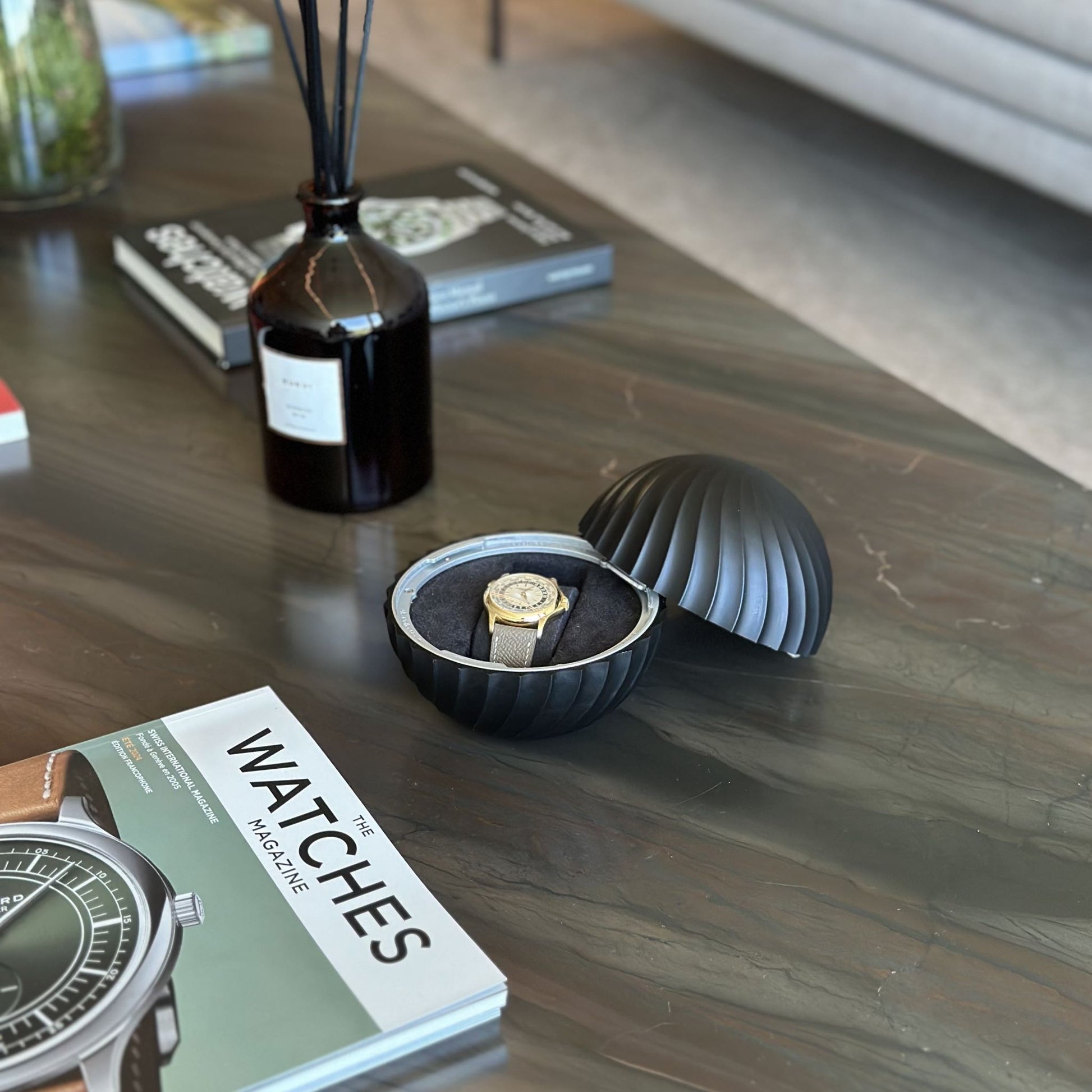 Open Obsidian Black ChronoSphere with a Swiss gold watch resting on a dark marble living room table with horology magazines.